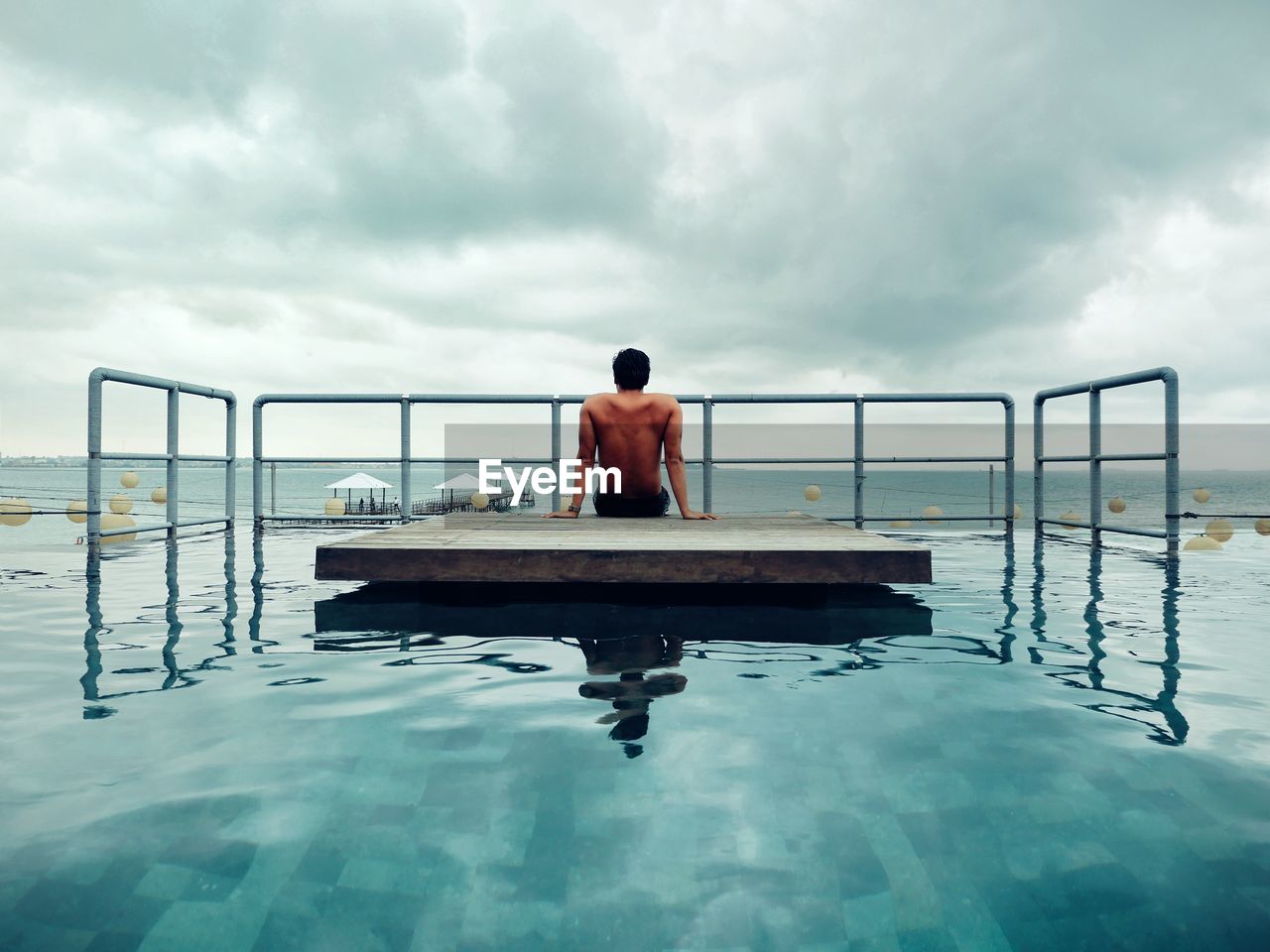 Shirtless man sitting on floating platform in swimming pool against sky