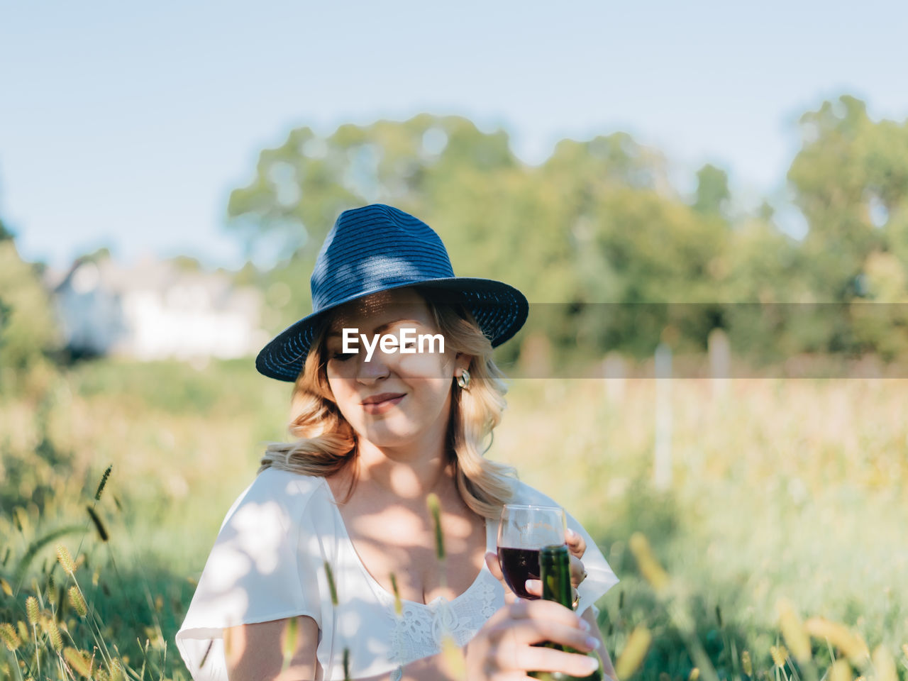 Young millennial woman outdoors in apple orchard having a glass of red wine with fedora hat on