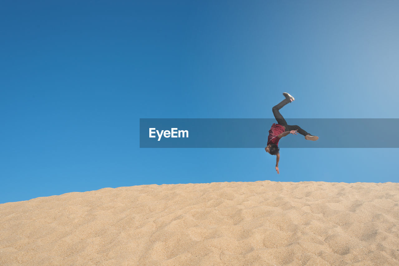 Full length of man jumping upside down on sand against clear blue sky