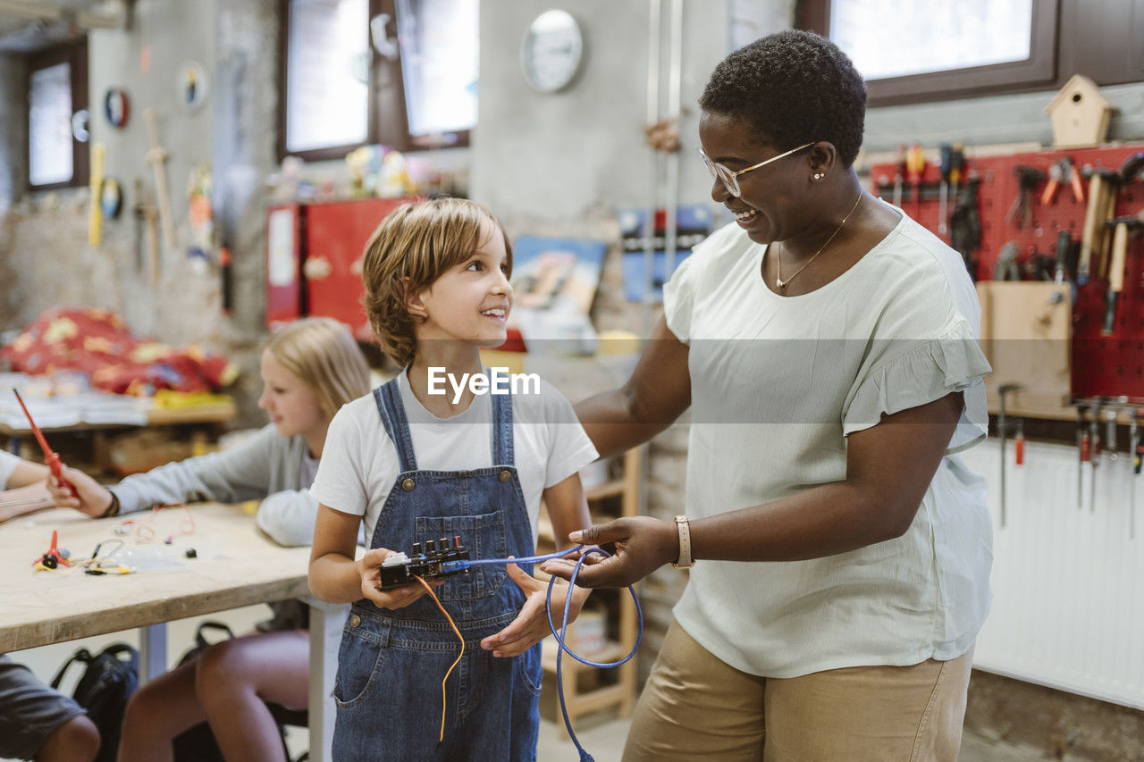 Smiling female teacher encouraging student holding electrical part at technology workshop in school