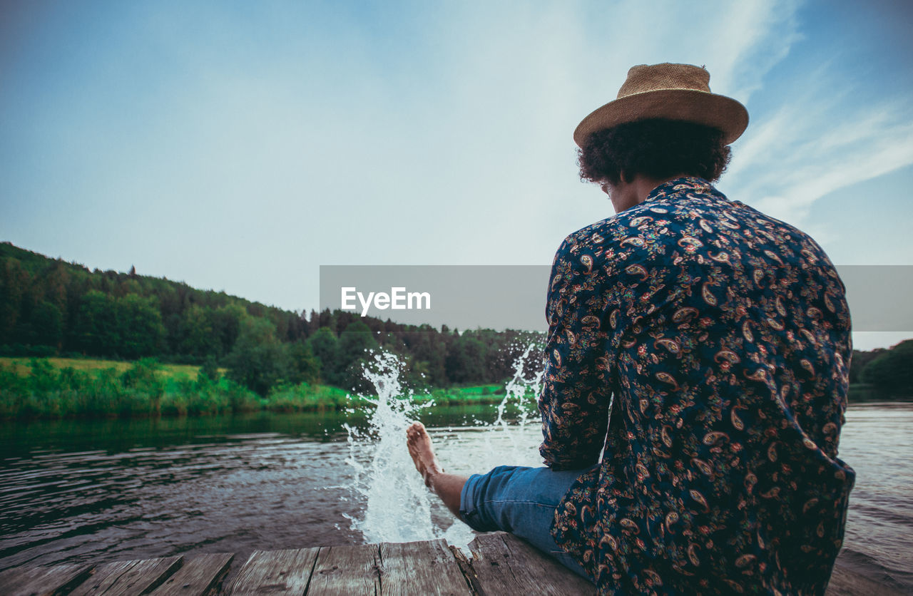 Rear view of man splashing water in lake while sitting on jetty against sky