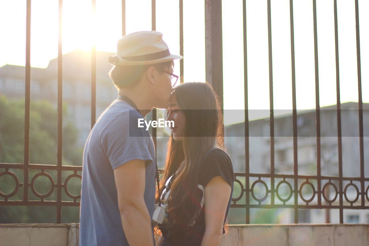 Young man kissing girlfriend while standing by fence