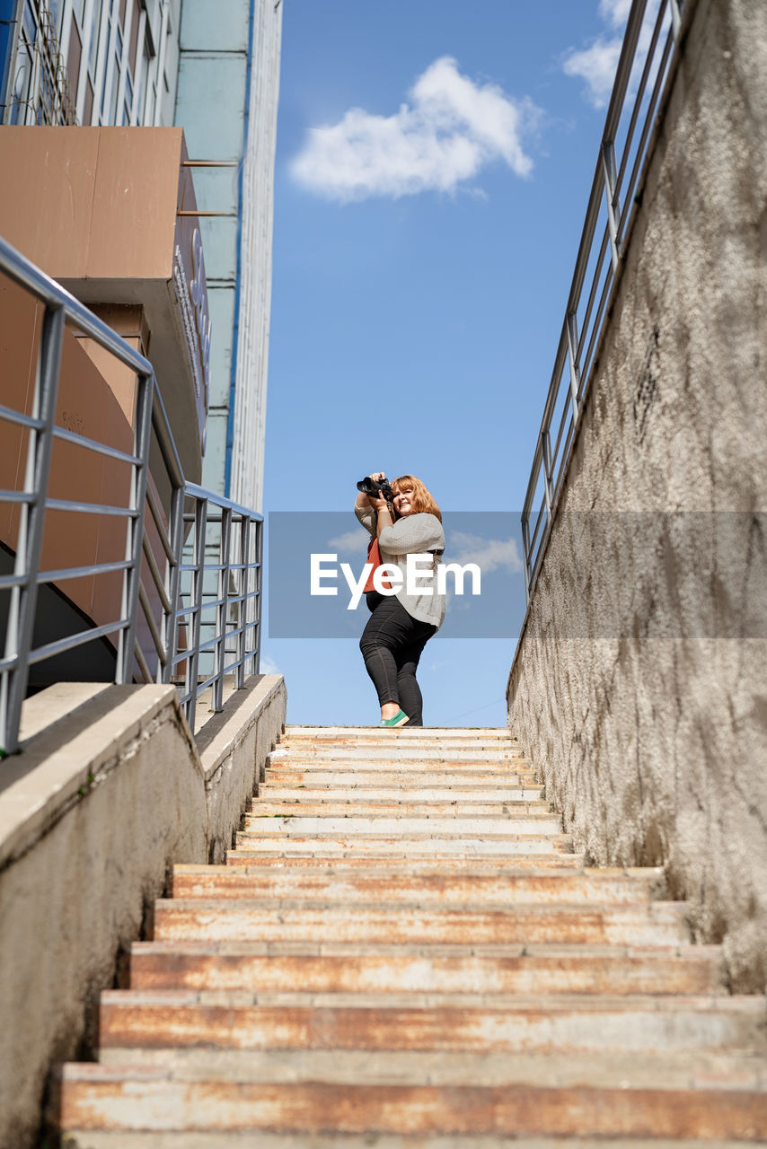 Smiling young woman holding camera standing against steps