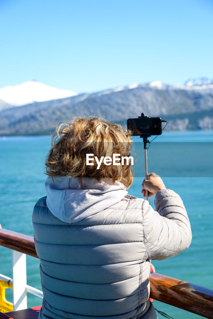 REAR VIEW OF WOMAN PHOTOGRAPHING AT SEA SHORE AGAINST SKY