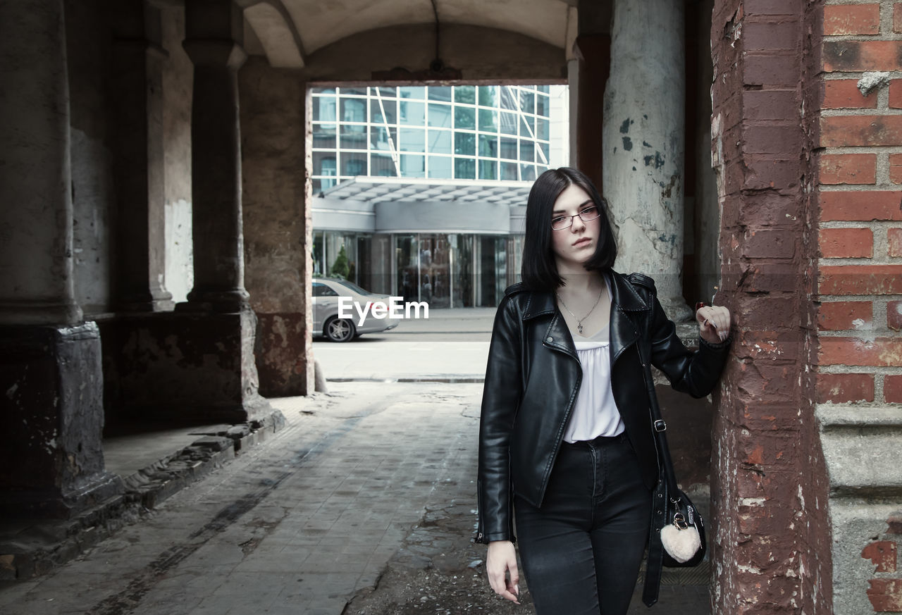 Sad young girl in black jacket and jeans stands in old arch on city street on summer day