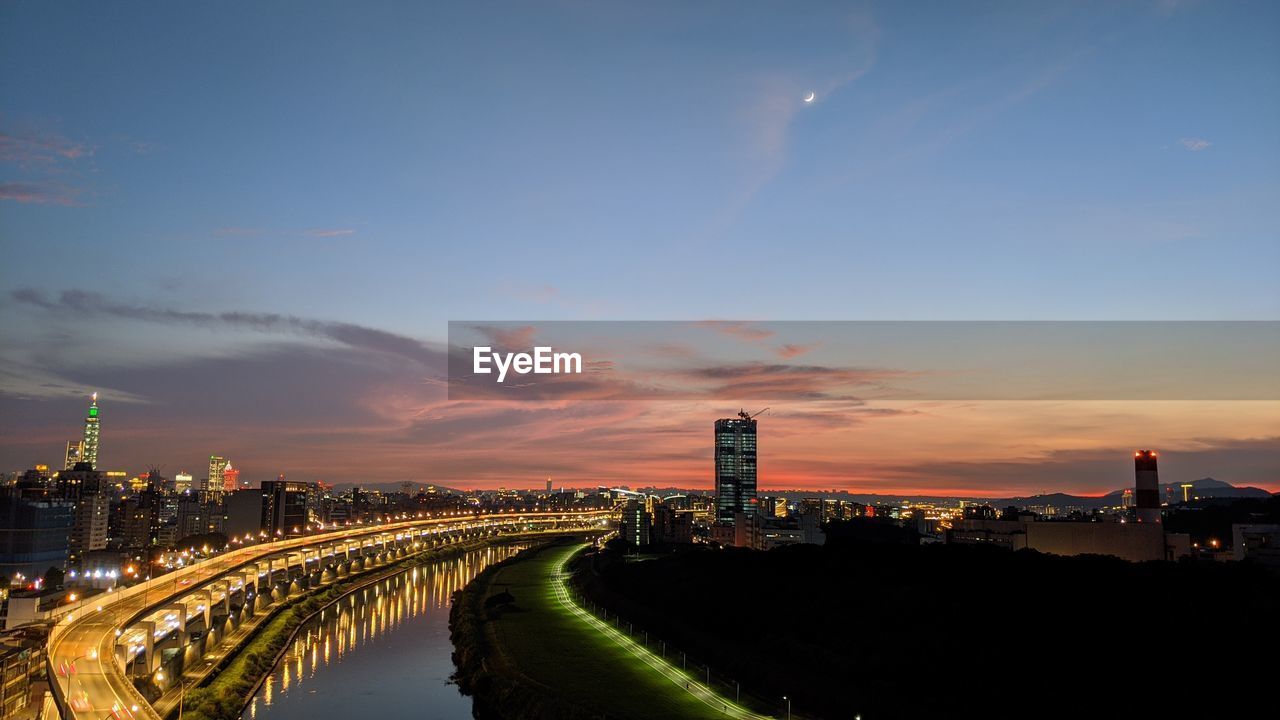 ILLUMINATED MODERN BUILDINGS BY RIVER AGAINST SKY DURING SUNSET