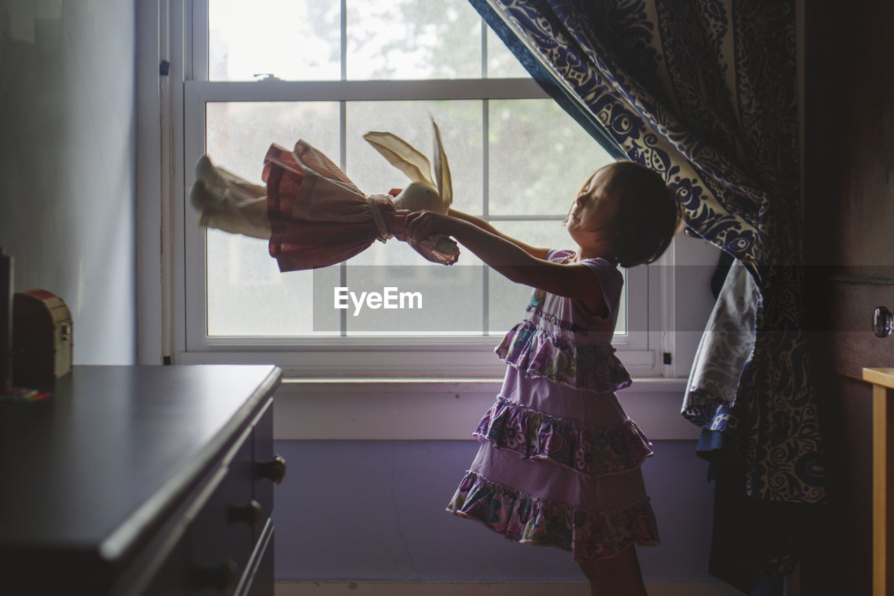 A cute little girl stands by window playing with a soft stuffed animal