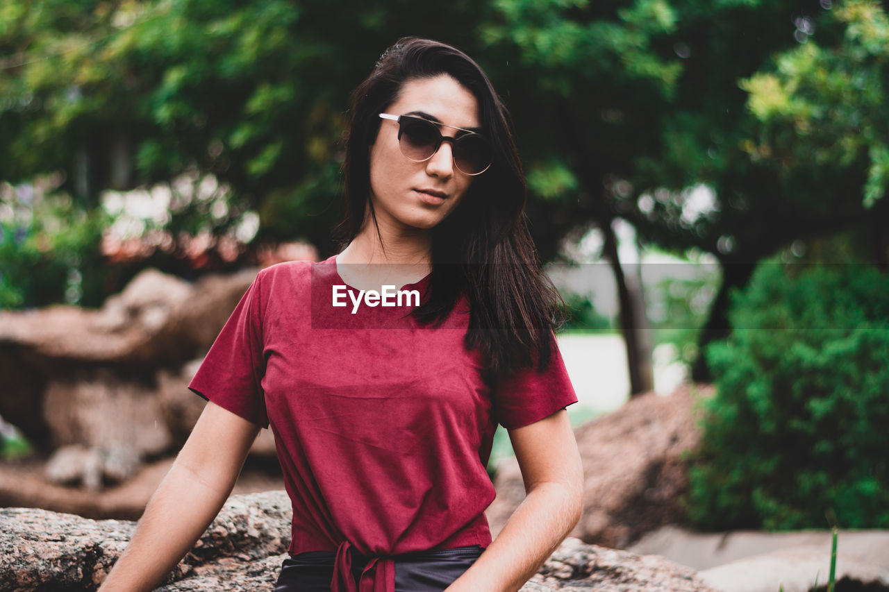 BEAUTIFUL YOUNG WOMAN WEARING SUNGLASSES STANDING AGAINST BLURRED BACKGROUND