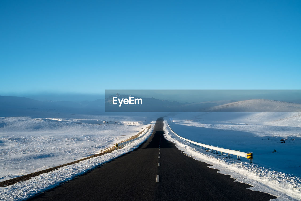 Empty road leading towards snowcapped mountains against clear blue sky