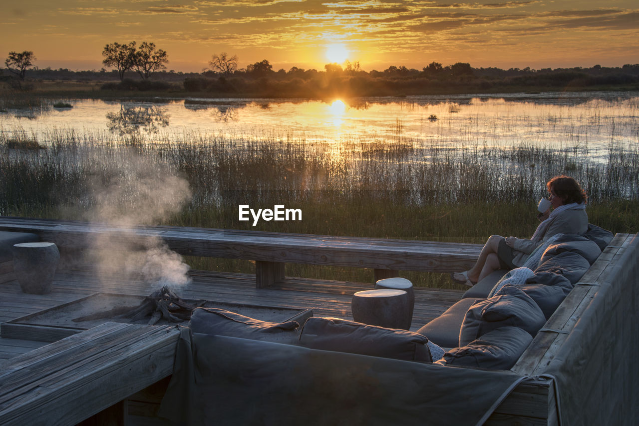 Woman drinking coffee on sofa against lake during sunset