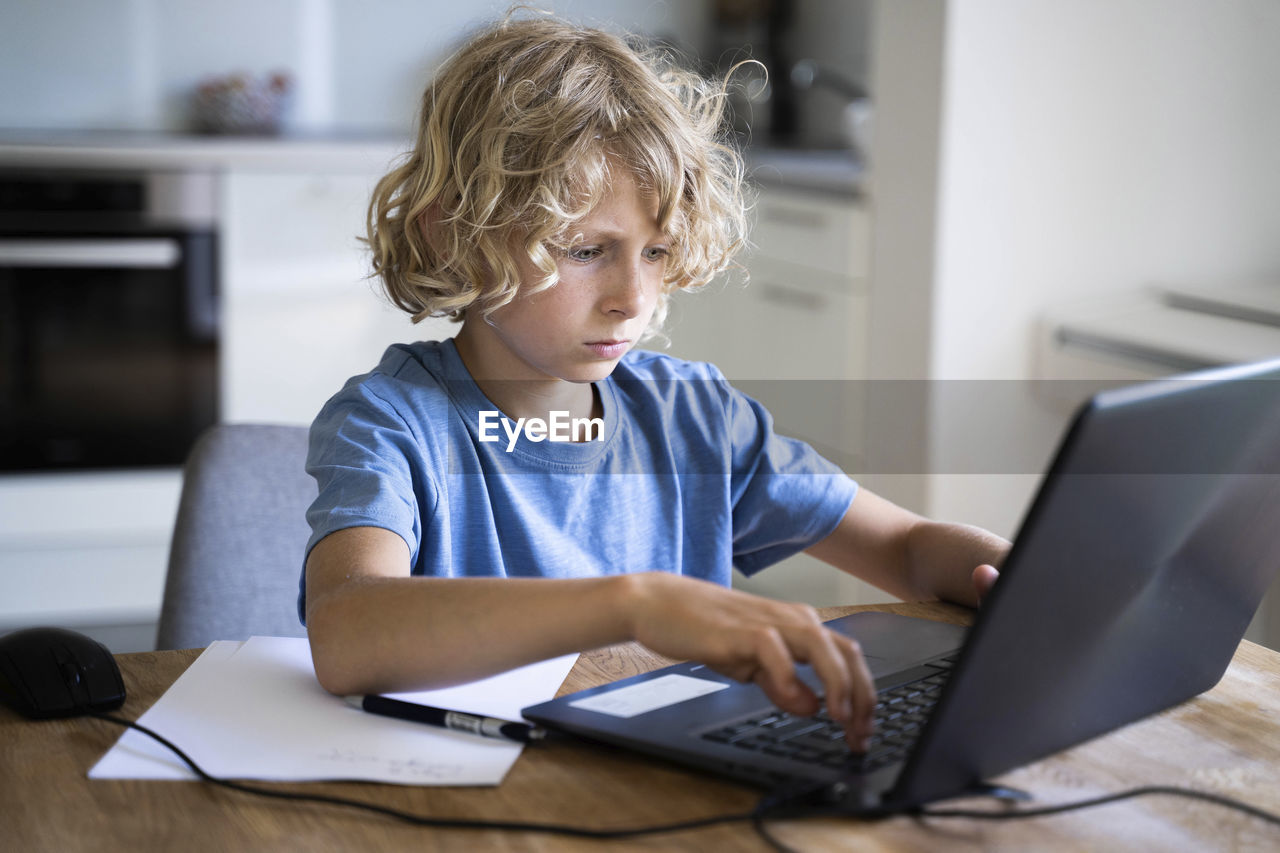 Boy with blond hair using laptop on table at home