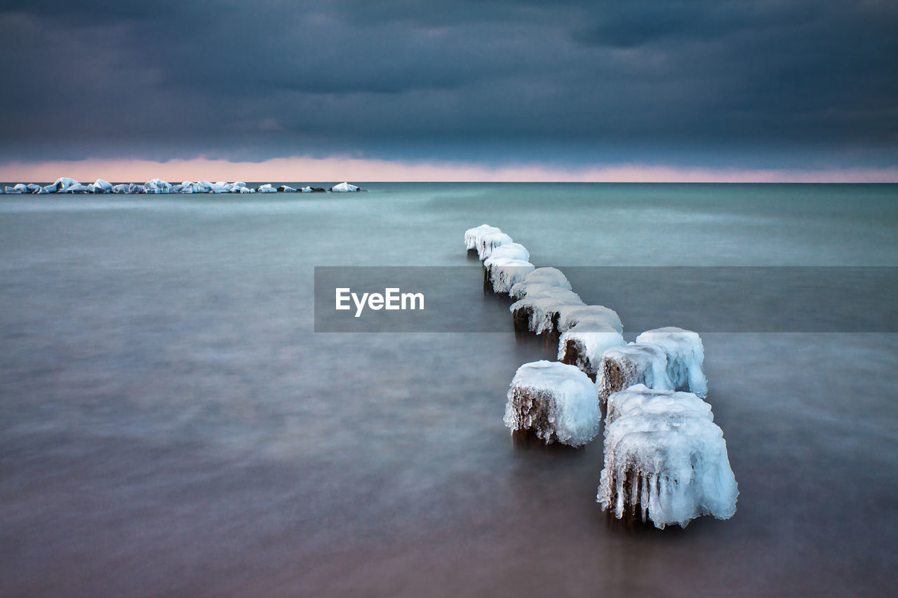 Frozen wooden posts in sea against cloudy sky