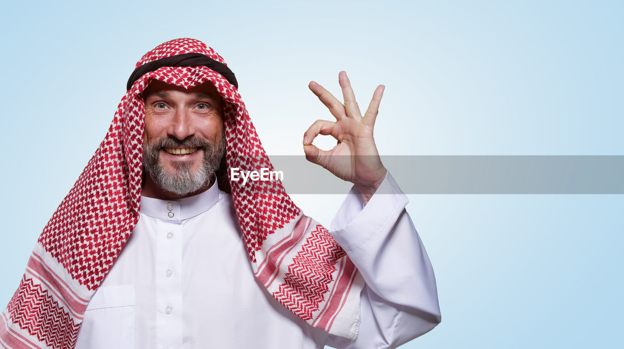 Cheerful and confident arab man smiles warmly and shows ok sign against light blue background