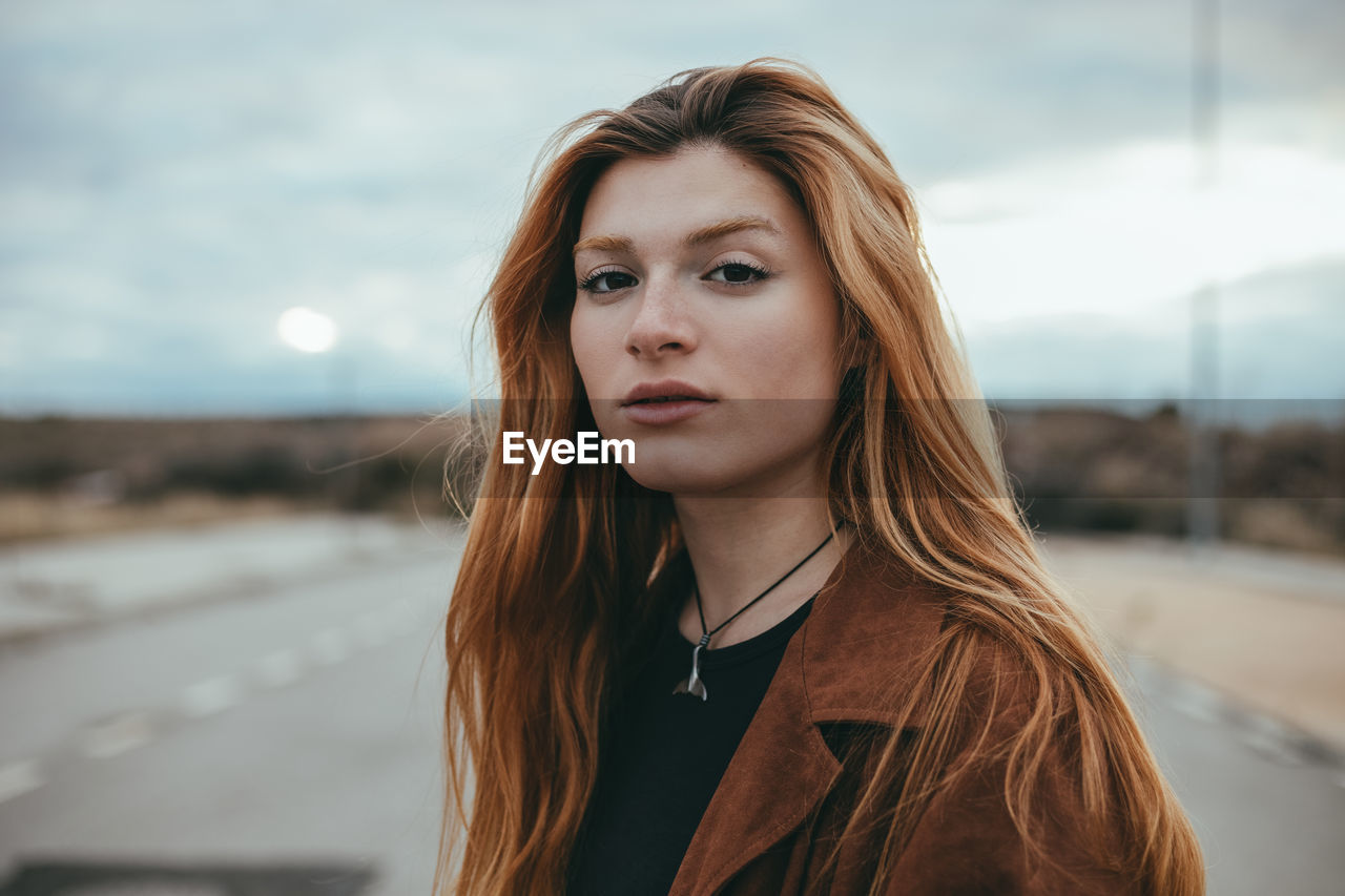 Confident young female with long ginger hair standing on street on cloudy day and looking at camera