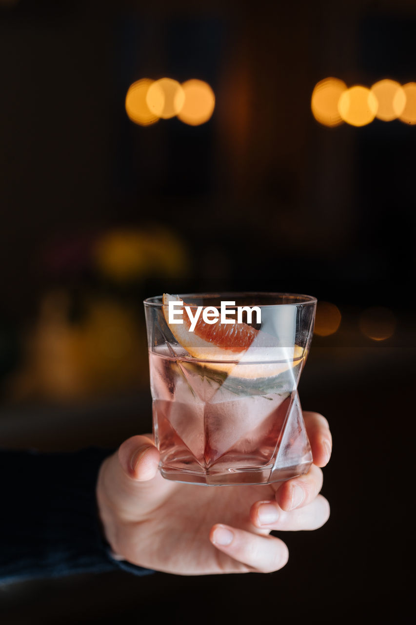 Cropped unrecognizable person hand holding glass with cold alcoholic cocktail with slice of grapefruit and ice cube placed on table against black background