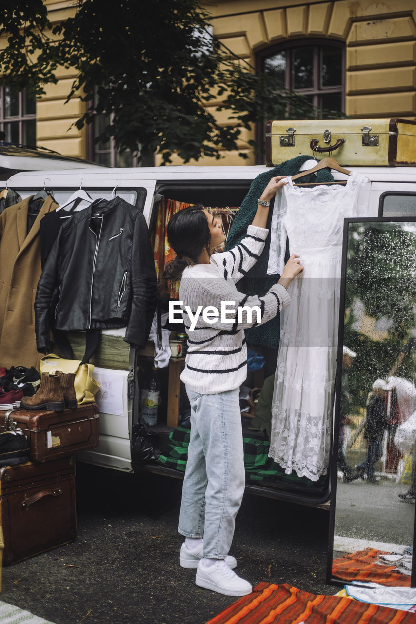 Side view full length of young female owner hanging dress during second hand sale at flea market