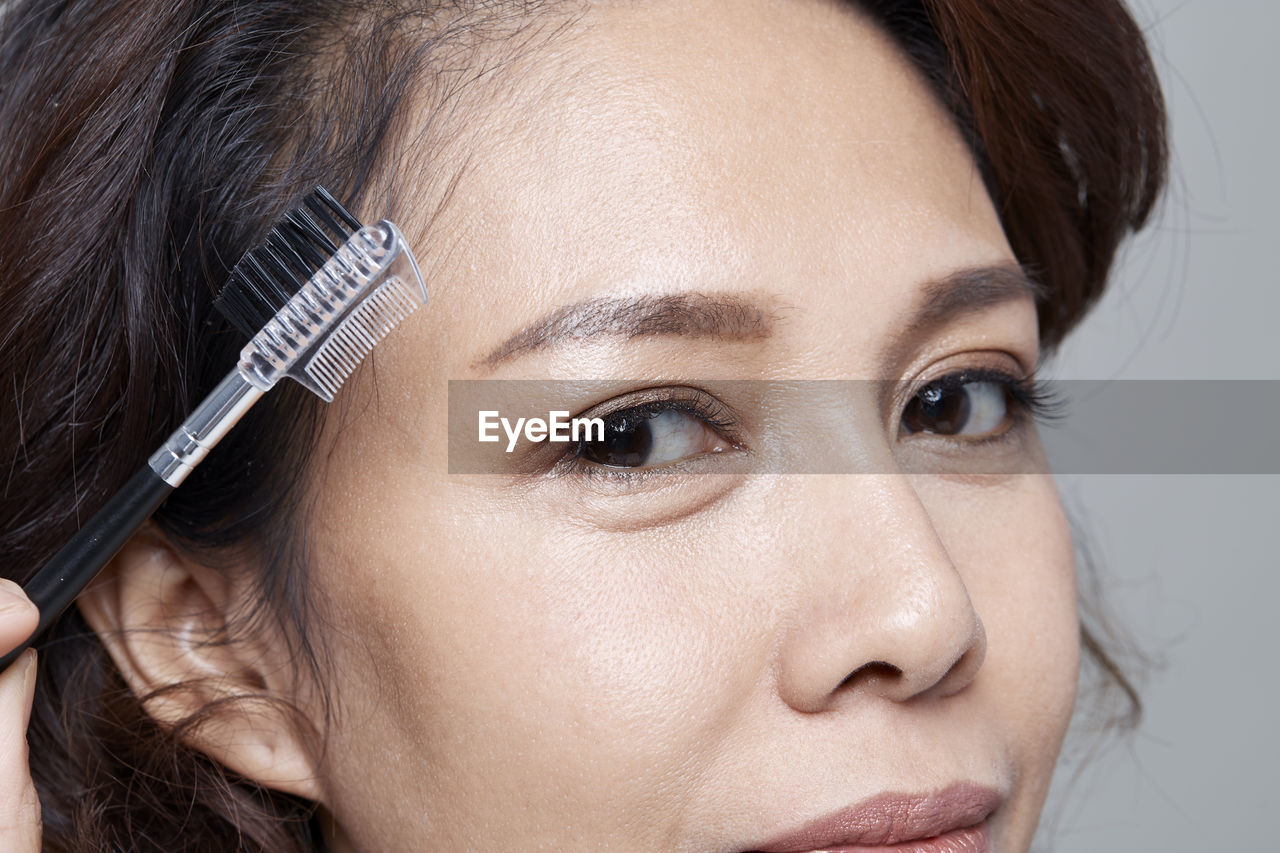 Close-up portrait of beautiful woman holding mascara applicator against gray background