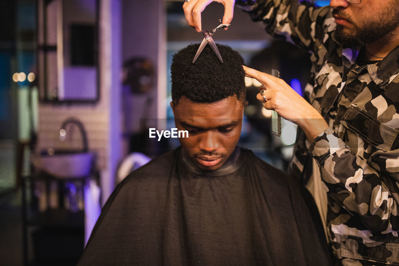 Crop focused barber with scissors standing near african american customer and making haircut in professional barbershop