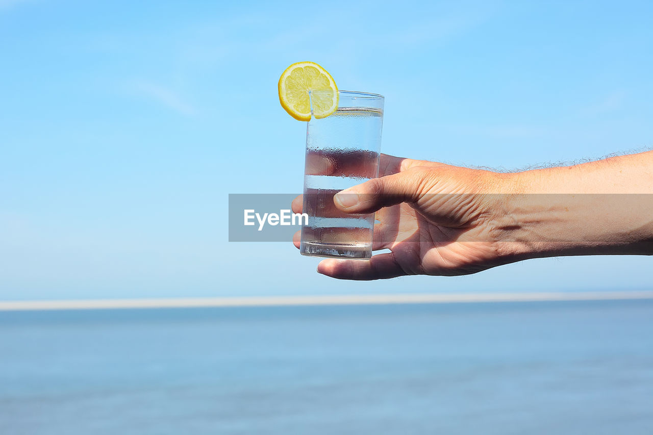 Close-up of hand holding glass of water against blue sky