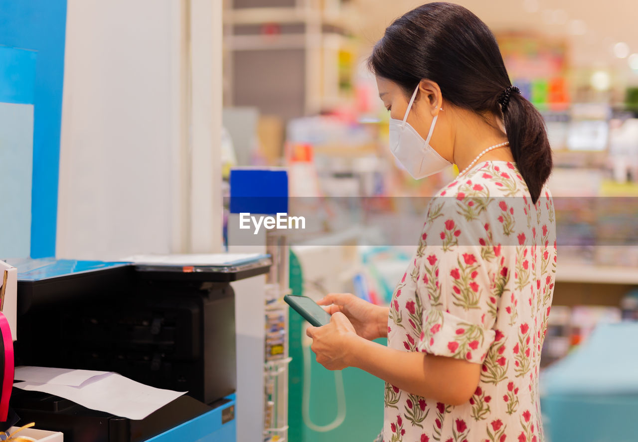 Cropped image of female in protective mask holding cell phone with printer to print the document.