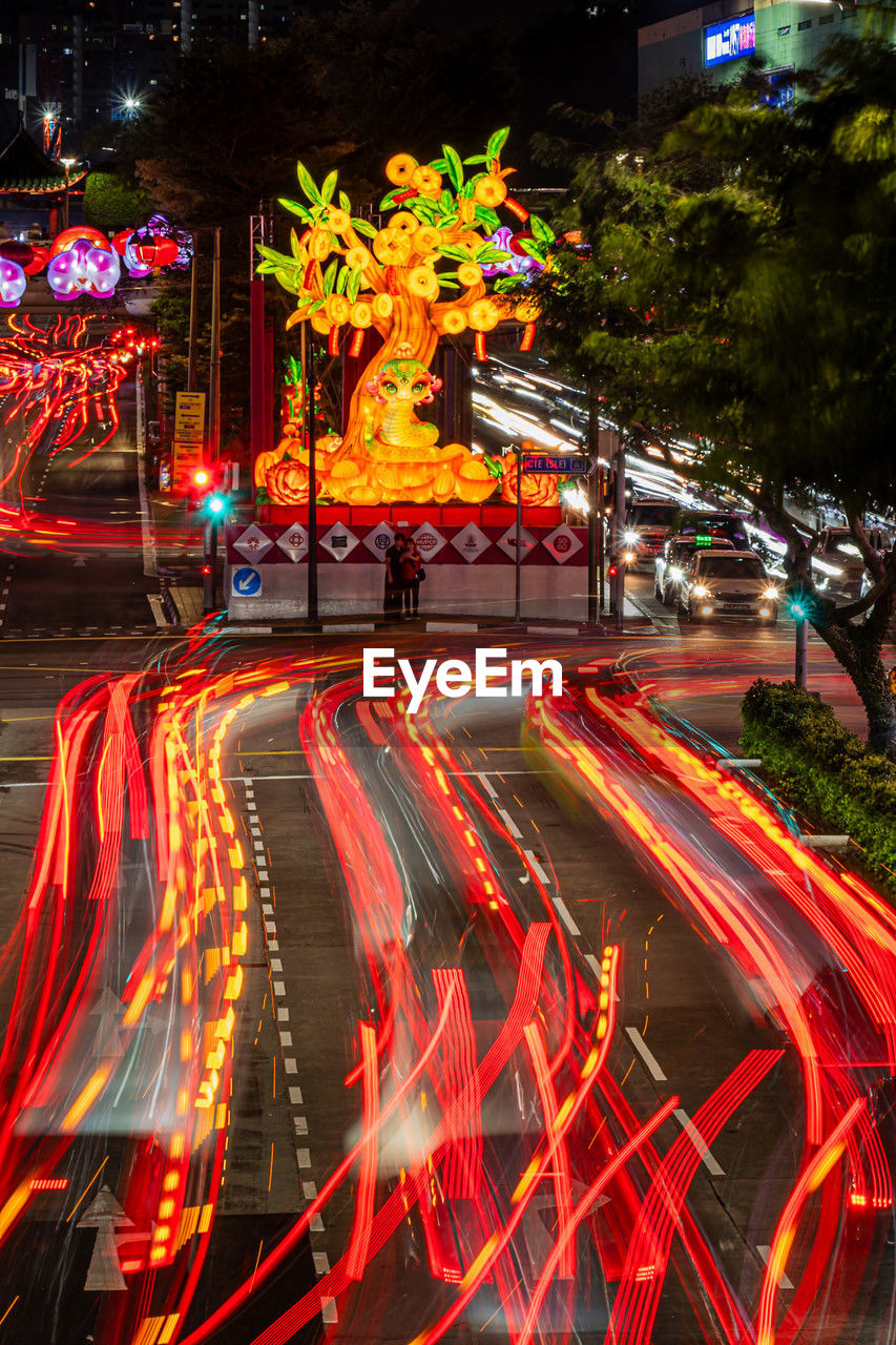illuminated, long exposure, night, light trail, motion, city, architecture, street, transportation, speed, road, blurred motion, city street, light, tree, no people, city life, plant, red, multi colored, traffic, lighting equipment, nature, mode of transportation, building exterior, outdoors, built structure, tail light, motor vehicle, evening, travel destinations, car, celebration