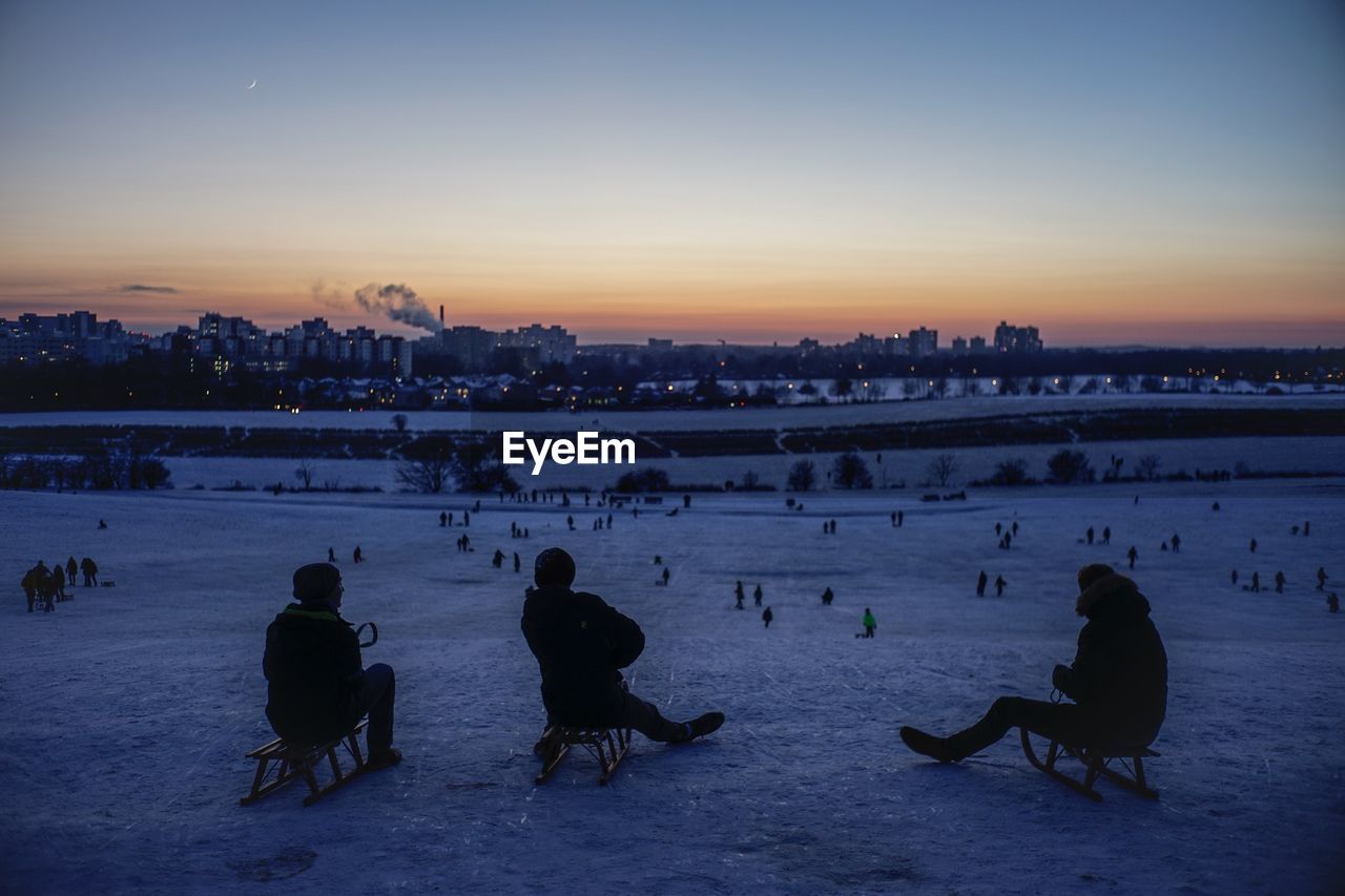 PEOPLE SITTING ON SNOW COVERED LANDSCAPE AGAINST SKY