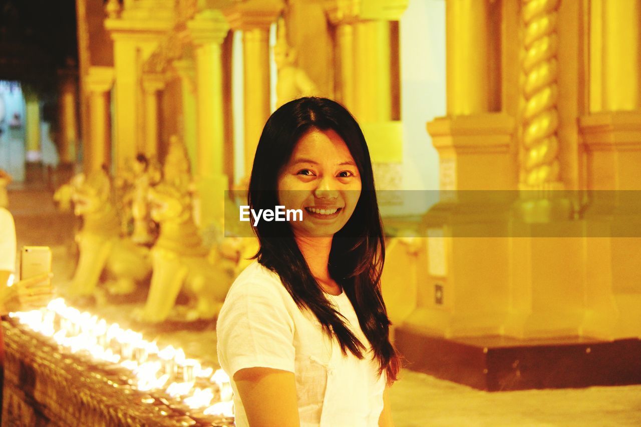 PORTRAIT OF SMILING YOUNG WOMAN AT ILLUMINATED TEMPLE