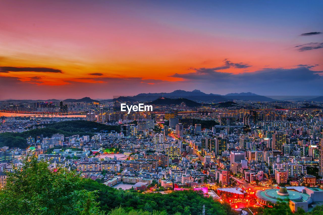 HIGH ANGLE VIEW OF ILLUMINATED CITY AGAINST SKY AT SUNSET