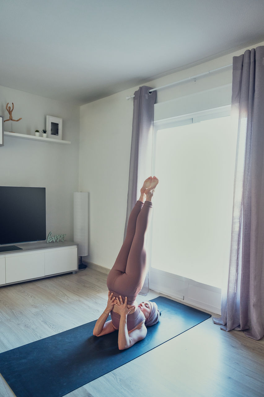 Young barefoot female in sportswear standing in salamba sarvangasana pose while practicing yoga on mat in house room