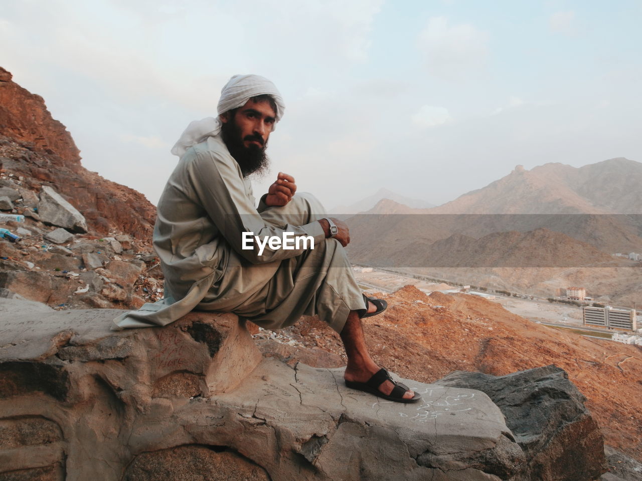 Side view of young man in traditional clothing sitting on rock against sky