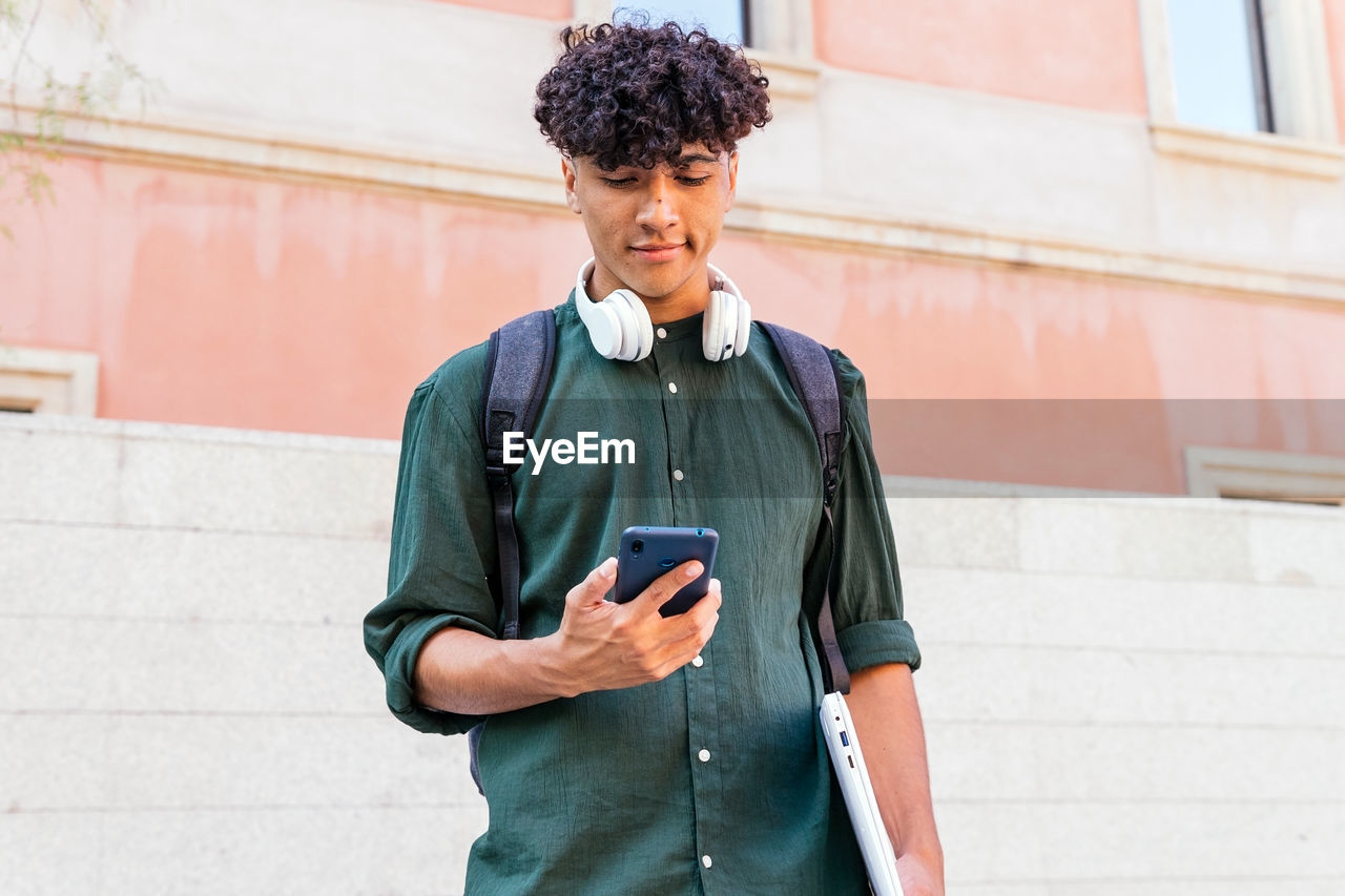 Ethnic man with headphones looking at screen of modern mobile phone on the street