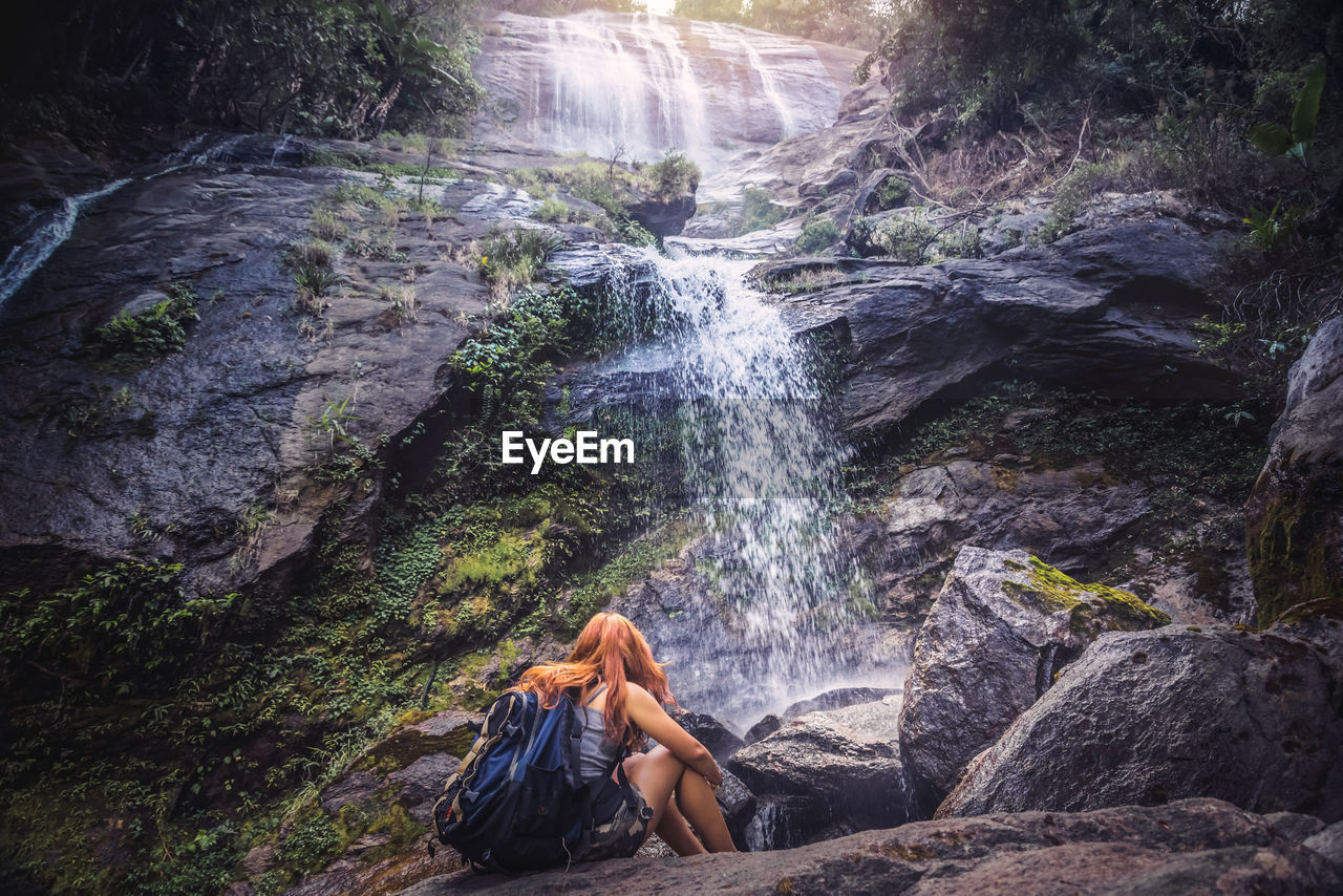 Rear view of woman with sitting on rock in forest against waterfall