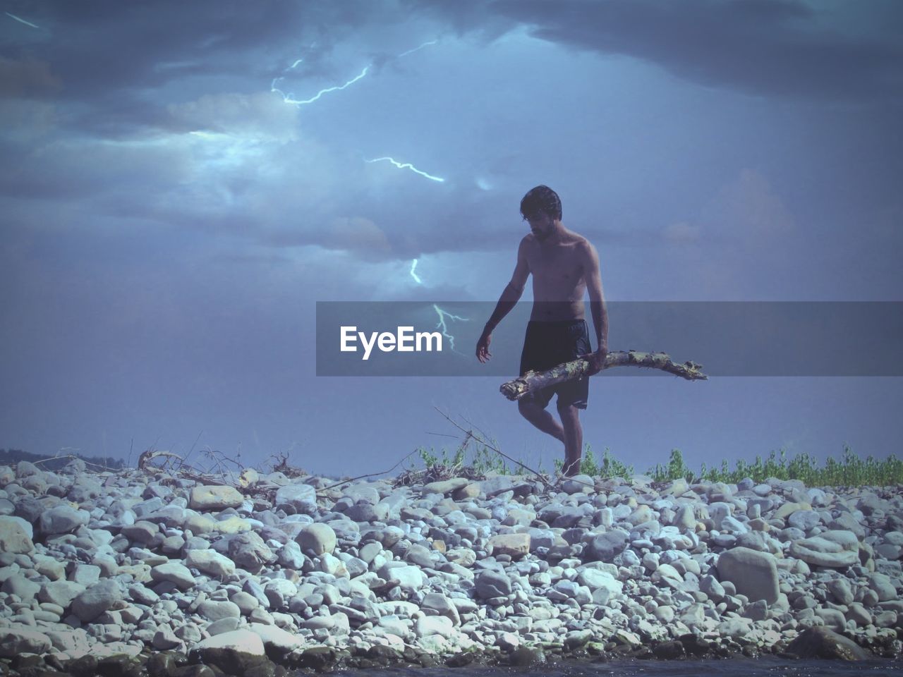 Full length of shirtless man carrying log on rocks against cloudy sky