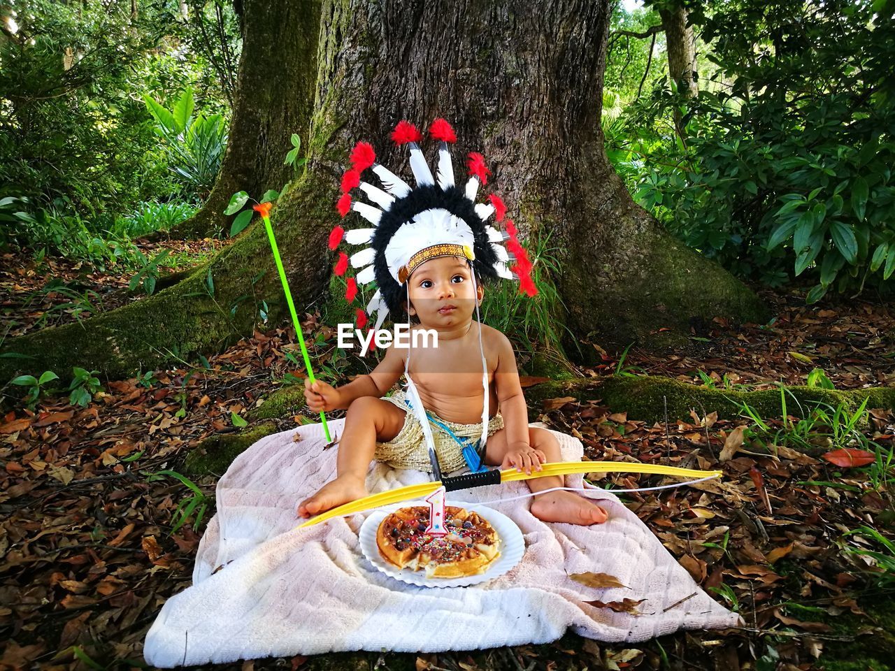 Baby boy wearing traditional headdress while sitting with birthday cake against tree in forest
