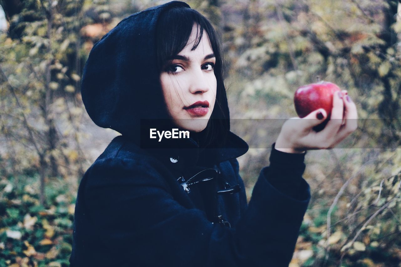 Portrait of young woman holding red apple while standing at park