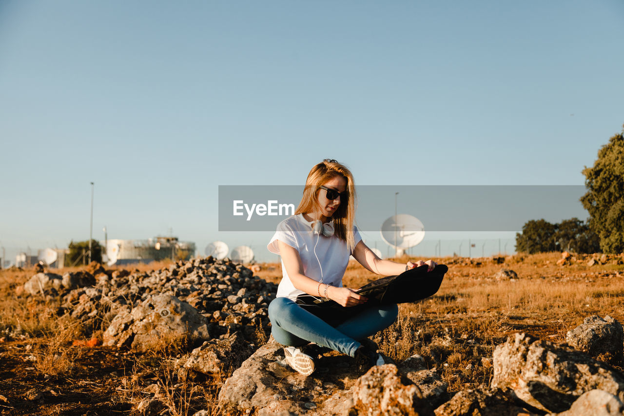 YOUNG WOMAN SITTING ON FIELD AGAINST SKY