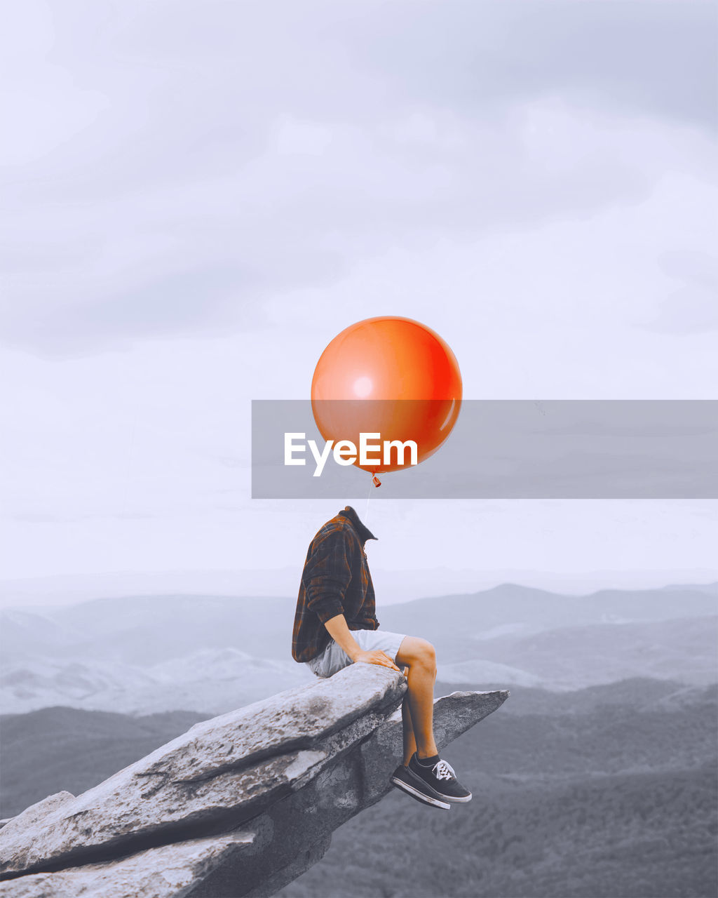 Digital composite image of decapitated man with balloon sitting on rock against sky