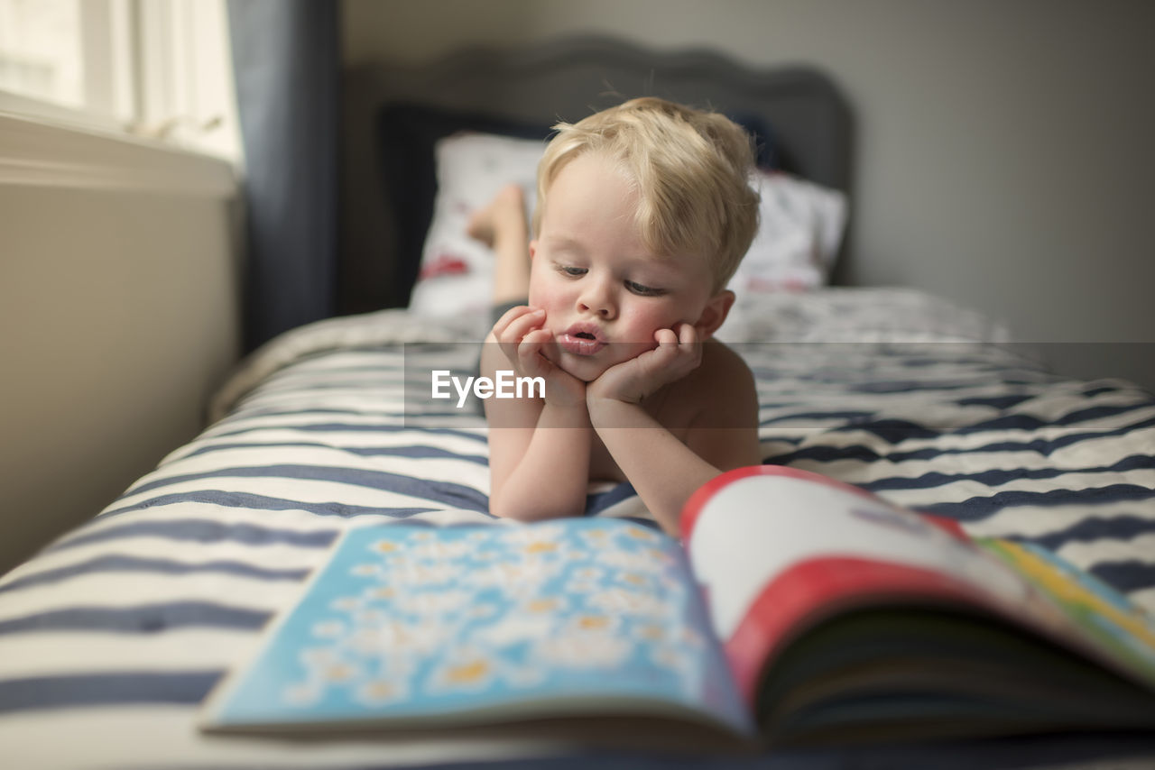 Boy with hands on chin reading picture book while lying on bed at home