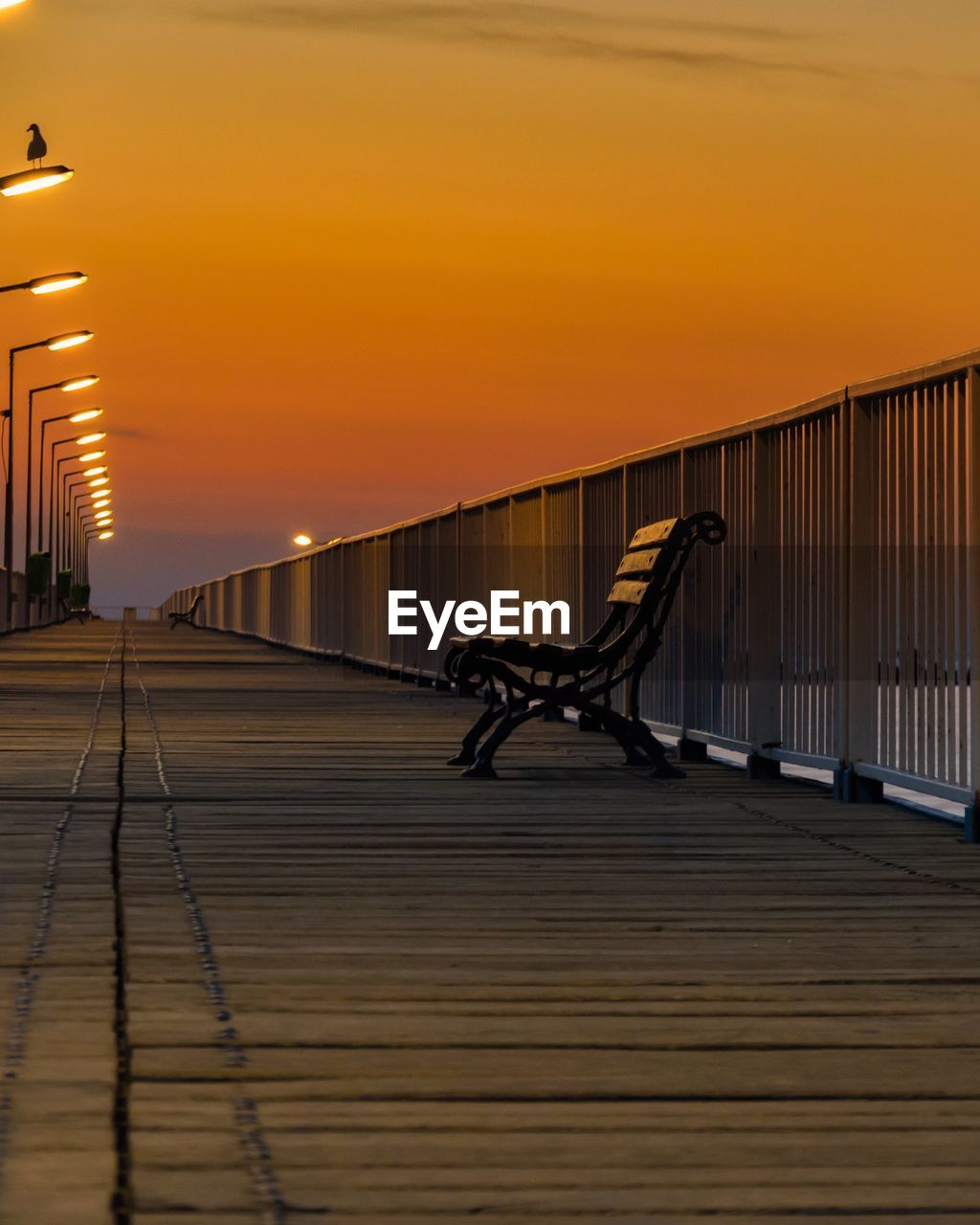 Empty bench on bridge against sky during sunset