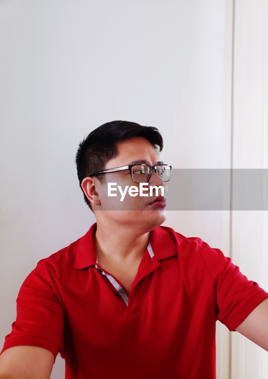 PORTRAIT OF YOUNG MAN WEARING EYEGLASSES AGAINST WALL