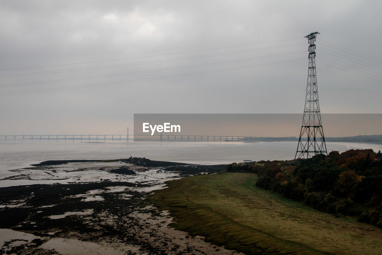 ELECTRICITY PYLONS ON FIELD BY SEA AGAINST SKY