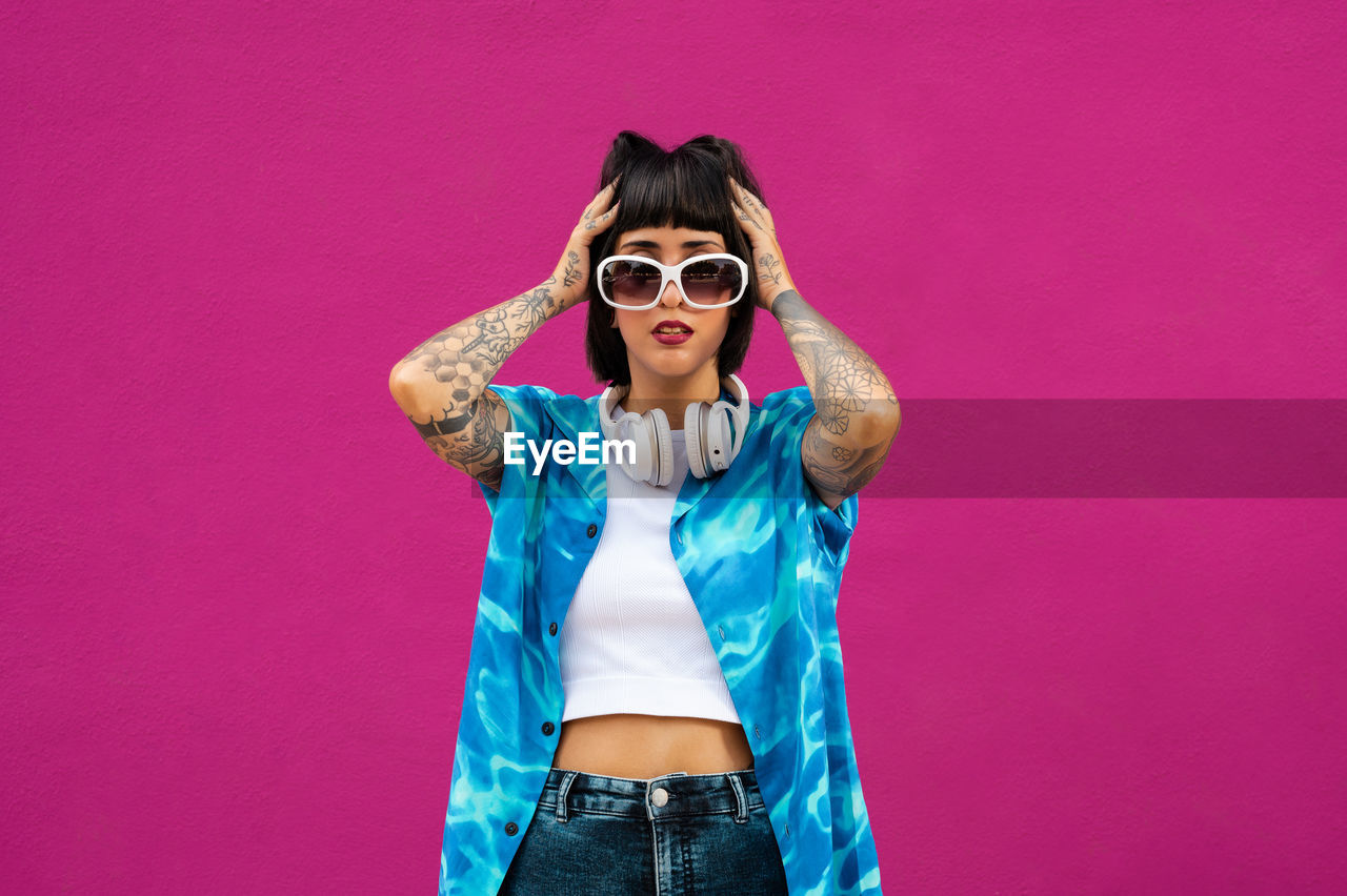 fashion, sunglasses, glasses, one person, colored background, pink, portrait, adult, studio shot, young adult, women, front view, fun, clothing, looking at camera, casual clothing, pink background, standing, indoors, hairstyle, arts culture and entertainment, three quarter length, emotion, photo shoot, cool attitude, happiness, smiling, purple, copy space, waist up, lifestyles, humor, blue, female, gesturing, positive emotion, brown hair, individuality