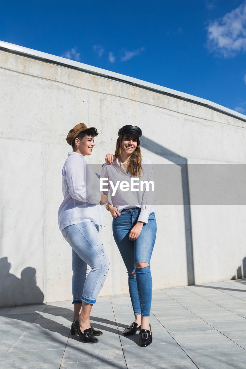 Full body of young positive female friends in trendy outfits and hats standing on walkway near gray wall in sunny day under blue sky