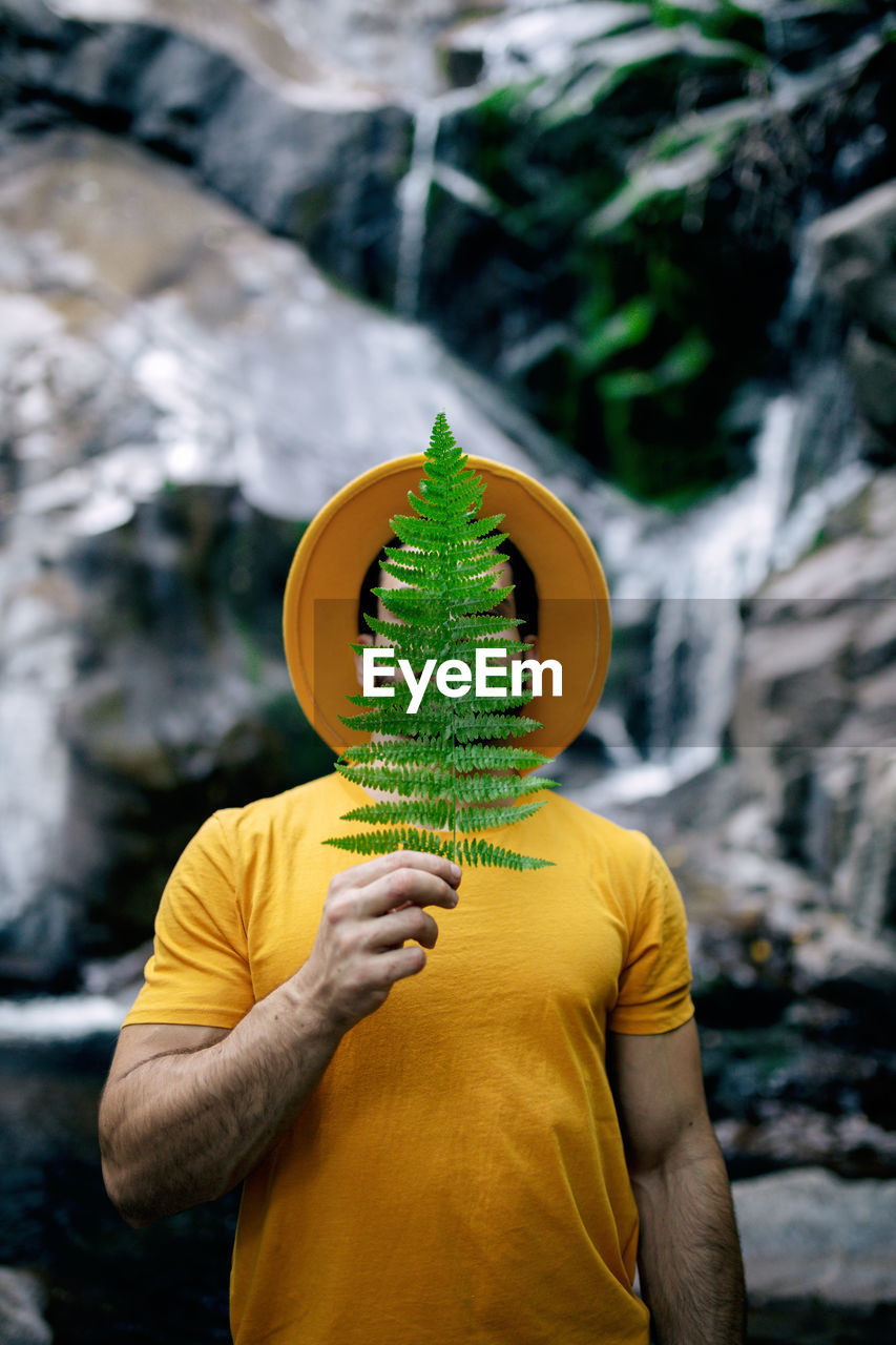 Peaceful male traveler in yellow clothes standing with green fern leaf on face and enjoying nature on background of waterfall in forest