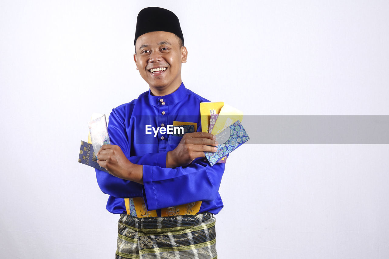 Portrait of smiling young man holding envelopes during traditional festival against white background