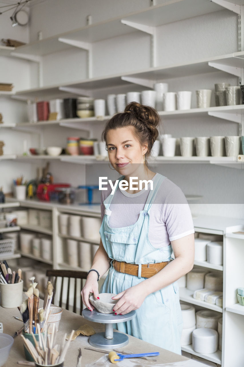 Portrait of smiling young potter molding shape on clay while standing by workbench at store