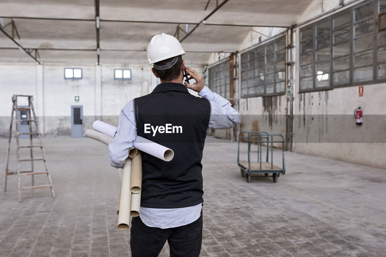 Male architect holding cardboards talking over mobile phone while standing in building