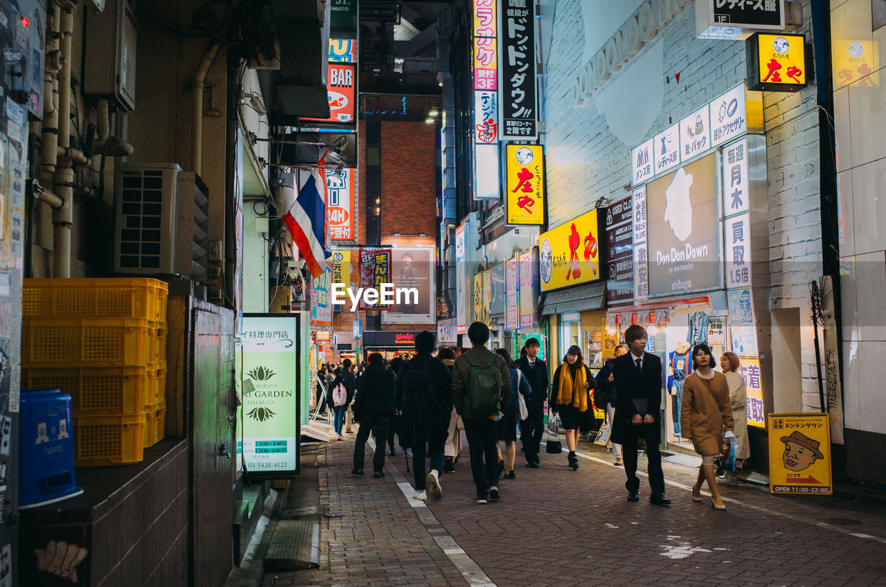 PEOPLE WALKING ON CITY STREET AMIDST BUILDINGS IN ILLUMINATED TOWN