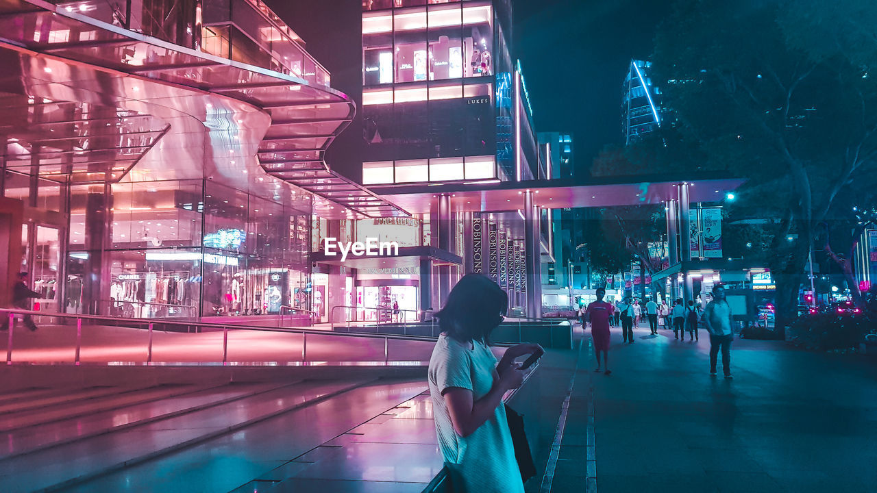 Side view of woman using phone by illuminated building in city at night