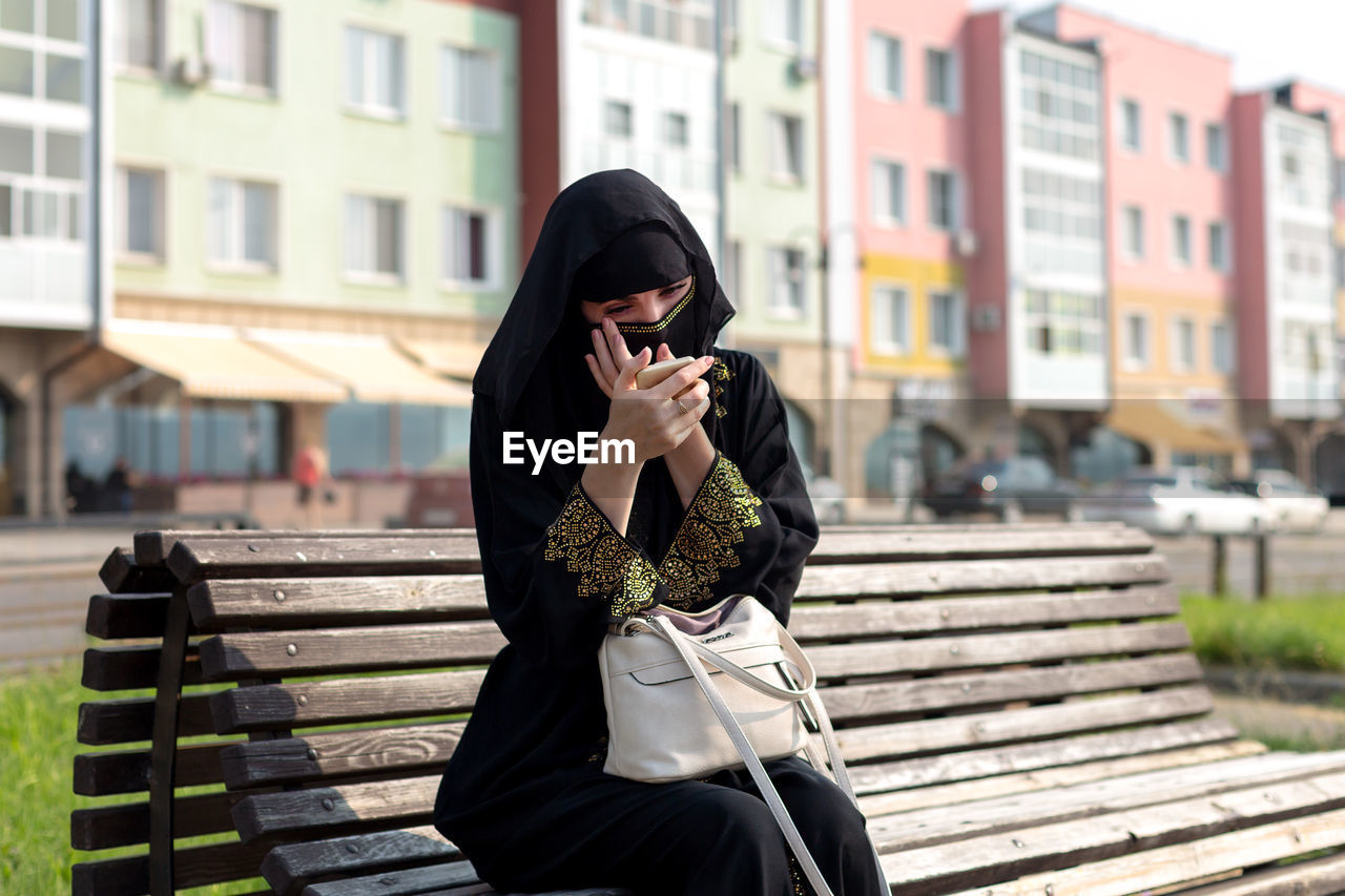 A muslim woman is resting on a bench in the park, tidying herself up looking in a mirror.